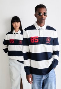 Marine- og hvitstripete langermede polo-skjorter med rød brodert nummer "85" og logo; modell med solbriller; avslappet denim-skjørt og jeans.