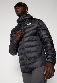 Hombre con chaqueta acolchada negra con capucha y gafas de sol, ajustando la cremallera de la chaqueta, de pie frente a un fondo claro y sencillo.