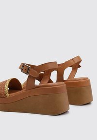 Brown platform sandals with woven detailing, adjustable ankle strap, and a smooth matte finish. Features a thick, light brown wedge sole.