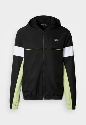 Chaqueta con capucha negra con cremallera y mangas largas. Rayas contrastantes en blanco y verde claro en las mangas. Logotipo de Lacoste en el lado izquierdo del pecho.