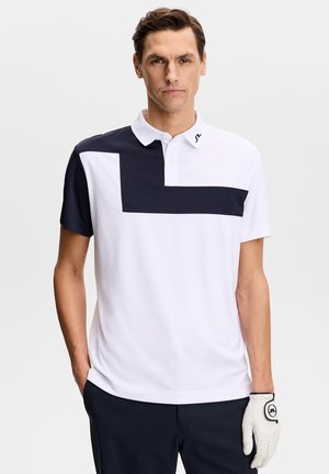 Uomo che indossa una polo bianca e blu navy con design geometrico, pantaloni blu navy e un guanto da golf bianco sulla mano destra, in piedi fermo.