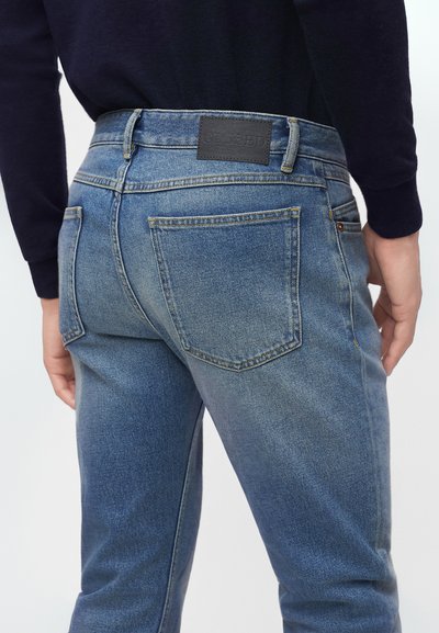 Personne portant un jean en denim bleu clair et un haut noir à manches longues, vue de dos de la taille jusqu'à mi-cuisse, avec des poches arrière visibles et une étiquette.