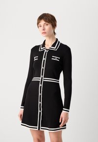 Robe noire à manches longues, avec des accents rayés blancs, un col et une fermeture à boutons. Dispose de deux poches avant avec des détails décoratifs.
