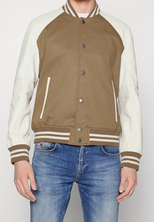 Homme portant une veste universitaire marron et blanche avec poignets et col en côtes rayées, associée à un jean bleu, mains détendues le long du corps.