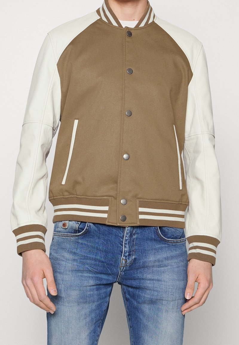 Homme portant une veste universitaire marron et blanche avec poignets et col en côtes rayées, associée à un jean bleu, mains détendues le long du corps.