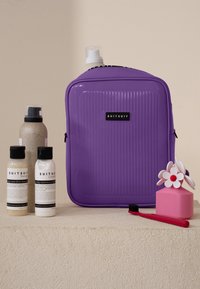 Paarse textuurkoffer met zwarte branding, drie flessen (crème en beige) met labels, een roze container in de vorm van een cadeau, en een rode tandenborstel.