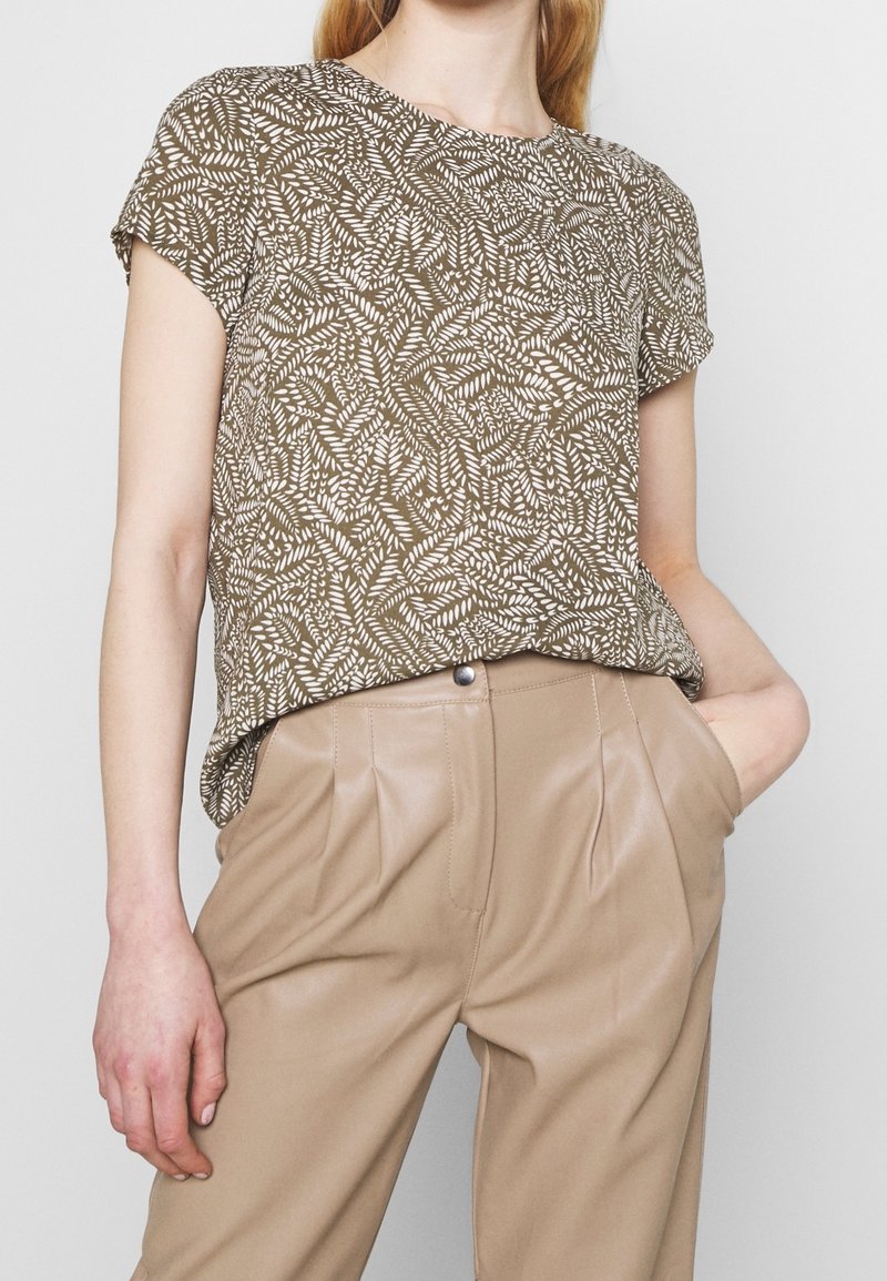 Femme portant un haut marron à manches courtes avec un motif de feuilles blanches rentré dans un pantalon beige plissé taille haute, une main dans la poche.