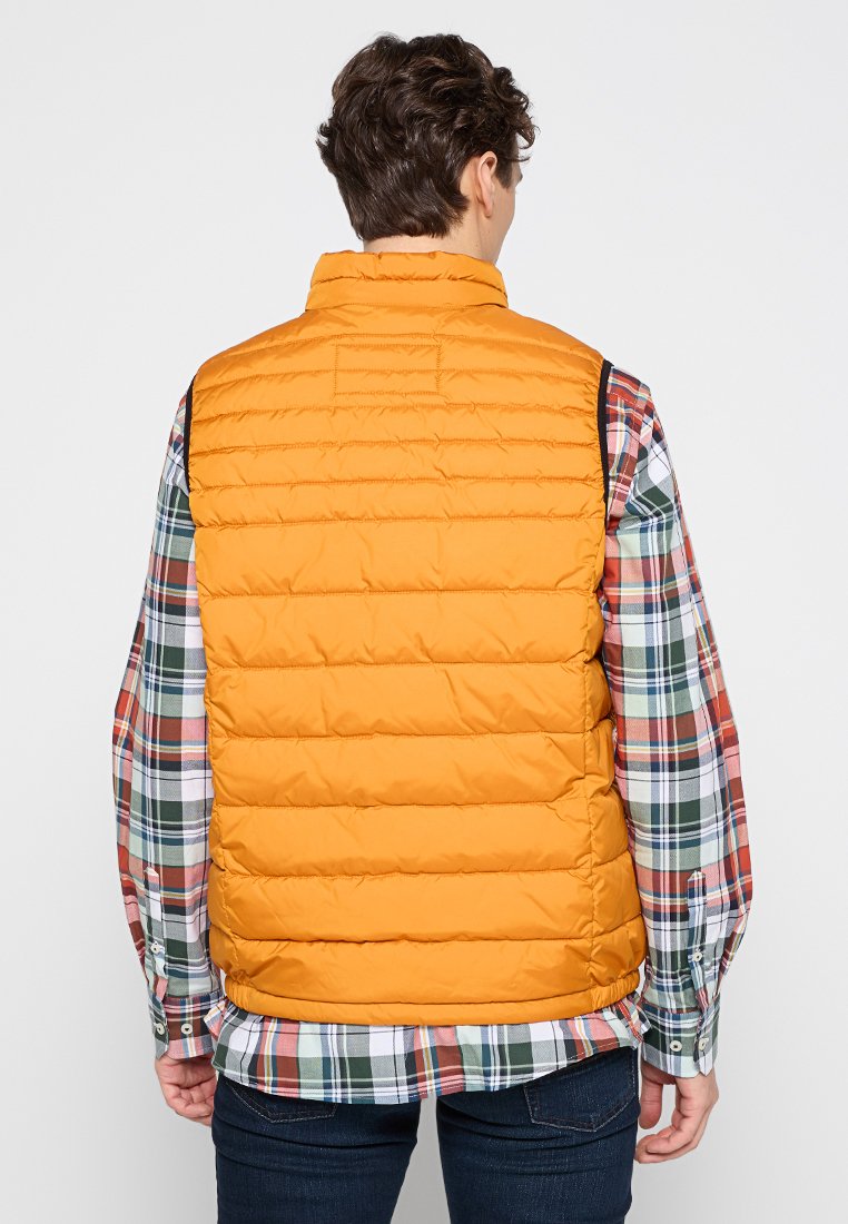 Gilet rembourré orange avec des coutures horizontales, porté sur une chemise à carreaux multicolore, avec un col haut et sans manches.
