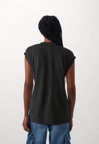 Osoba z długimi warkoczami, ubrana w bezrękawnik w kolorze czarnym oraz niebieskie spodnie cargo, odwrócona plecami na tle białego, gładkiego tła.