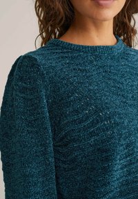 Femme portant un pull en tricot turquoise avec un motif texturé ondulé et un col rond, montrant le cou et une partie du visage.