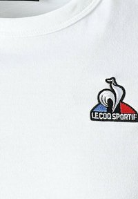 Camiseta blanca de algodón con cuello redondo, que presenta un logo de colores con elementos rojos, azules y negros bordados en el pecho.