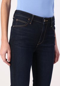 Jeans de denim azul oscuro con un diseño ajustado, herrajes de latón y costuras amarillas contrastantes. Bolsillos visibles en la parte frontal.