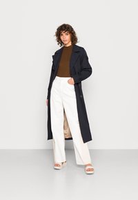 Trench coat azul-marinho com grandes lapelas, combinado com um topo ajustado castanho e calças largas brancas. O modelo usa sandálias de plataforma gordinhas em bege.