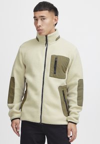Jakna s patentnim zatvaračem od flisa u boji off-white s maslinasto zelenim najlonskim zakrpama, crnim patentnim detaljima i visokim ovratnikom. Ima dva prednja džepa.