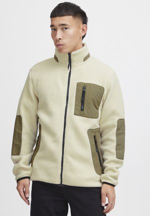 Veste en polaire zippée de couleur blanc cassé avec des patches en nylon vert olive, des accents de fermeture éclair noire et un col haut. Comprend deux poches avant.