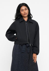 Jachetă croită neagră cu guler structurat, închidere cu fermoar și mâneci elastice. Purtați-o peste o rochie cu imprimeu de leopard întunecat.