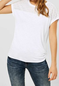 Bílý top s krátkým rukávem a strukturovaným síťovaným vzorem na horní části přední strany. Měkká tkanina, volný střih, s kulatým výstřihem a jemným řasením.