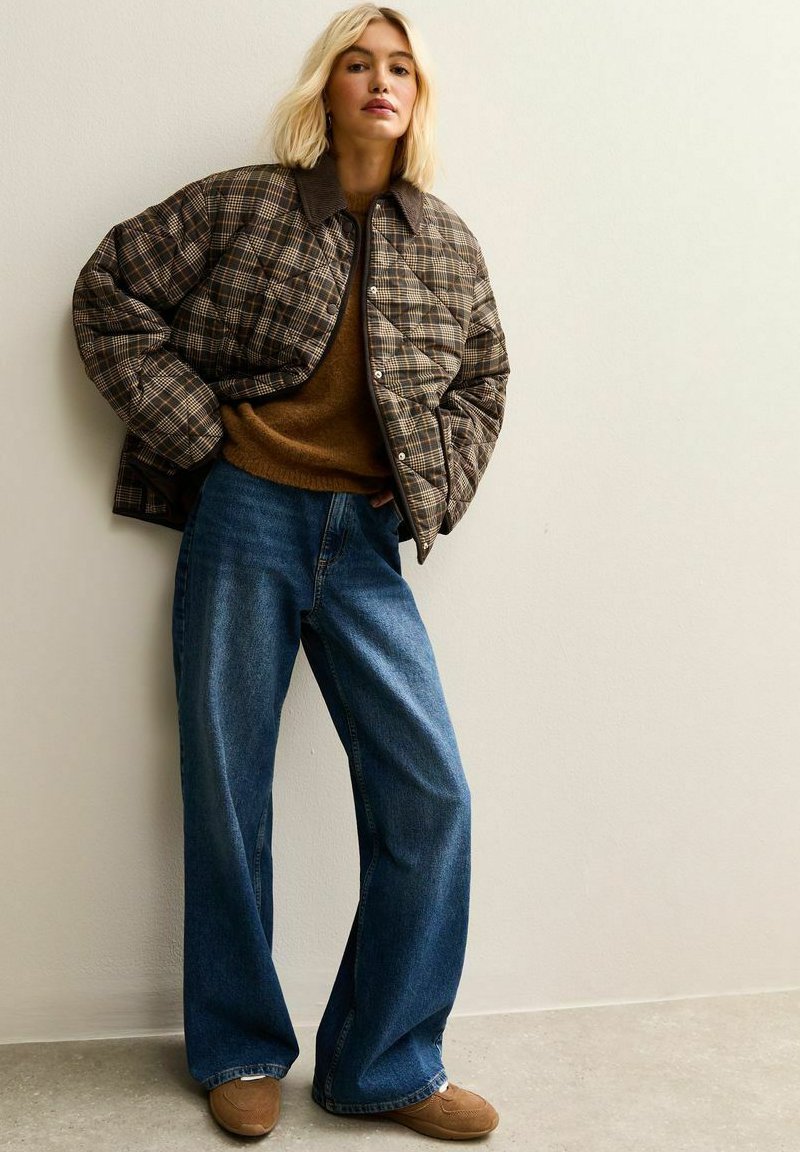 Blonde vrouw leunt tegen de muur in een oversized geruite jas, een bruine trui, wijde blauwe jeans en tan sneakers.