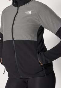 Persona che indossa una giacca in pile grigia e nera con zip a due toni, con il logo The North Face sul petto, mani nelle tasche laterali con zip.