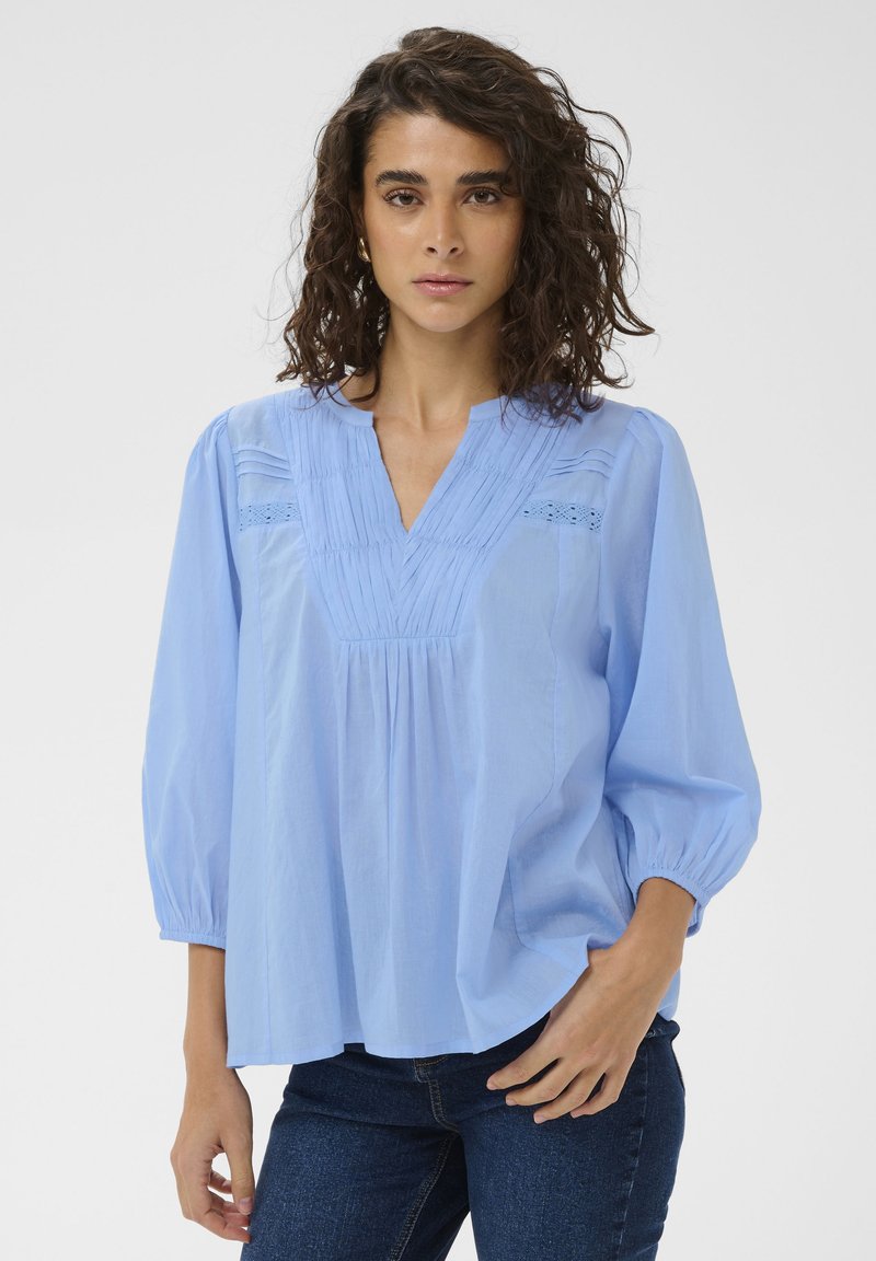 Blouse bleu clair à manches longues avec col en V, devant plissé et détails en dentelle œillets le long des épaules. Tissu doux, coupe décontractée.