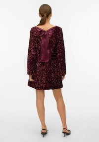 Bordeaux flonkerende jurk met een diepe V-rug en strikdetail. Lange mouwen, zachte textuur en een uitlopende silhouet met een lengte boven de knie.