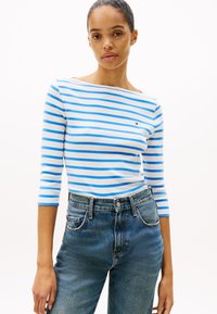 Ριγέ μακρυμάνικο μπλουζάκι σε λευκό και μπλε βαμβάκι με φαρδύ λαιμόκοψη, σε συνδυασμό με ψηλόμεσα μπλε τζιν παντελόνια.