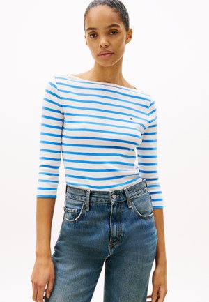 Prugasta dolga majica z dolgimi rokavi v beli in modri barvi iz bombaža s čolnastim ovratnikom, v kombinaciji z visokimi modrimi kavbojkami.