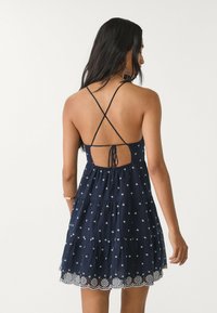 Granatowa, warstwowa sukienka letnia z białym haftem kwiatowym i regulowanymi skrzyżowanymi ramiączkami. Tył ma zawiązywane otwarcie z teksturowanym splotem.