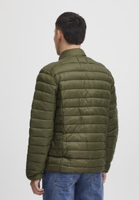 Olive green puffer jacket featuring quilted sections and a high collar, paired with blue denim jeans. Smooth texture, lightweight design.