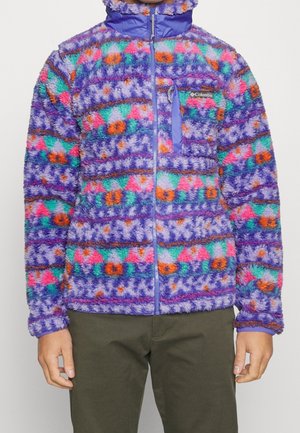 Persona che indossa una giacca in pile dai colori vivaci con motivi viola, arancione, rosa e teal, dotata di cerniera frontale e tasca sul petto, abbinata a pantaloni verde oliva.