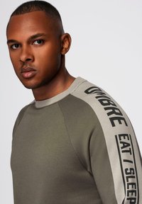 Sudadera de cuello redondo en verde oliva, con mangas raglán y texto negro en una banda crema a lo largo del brazo izquierdo. Material de mezcla de algodón suave.