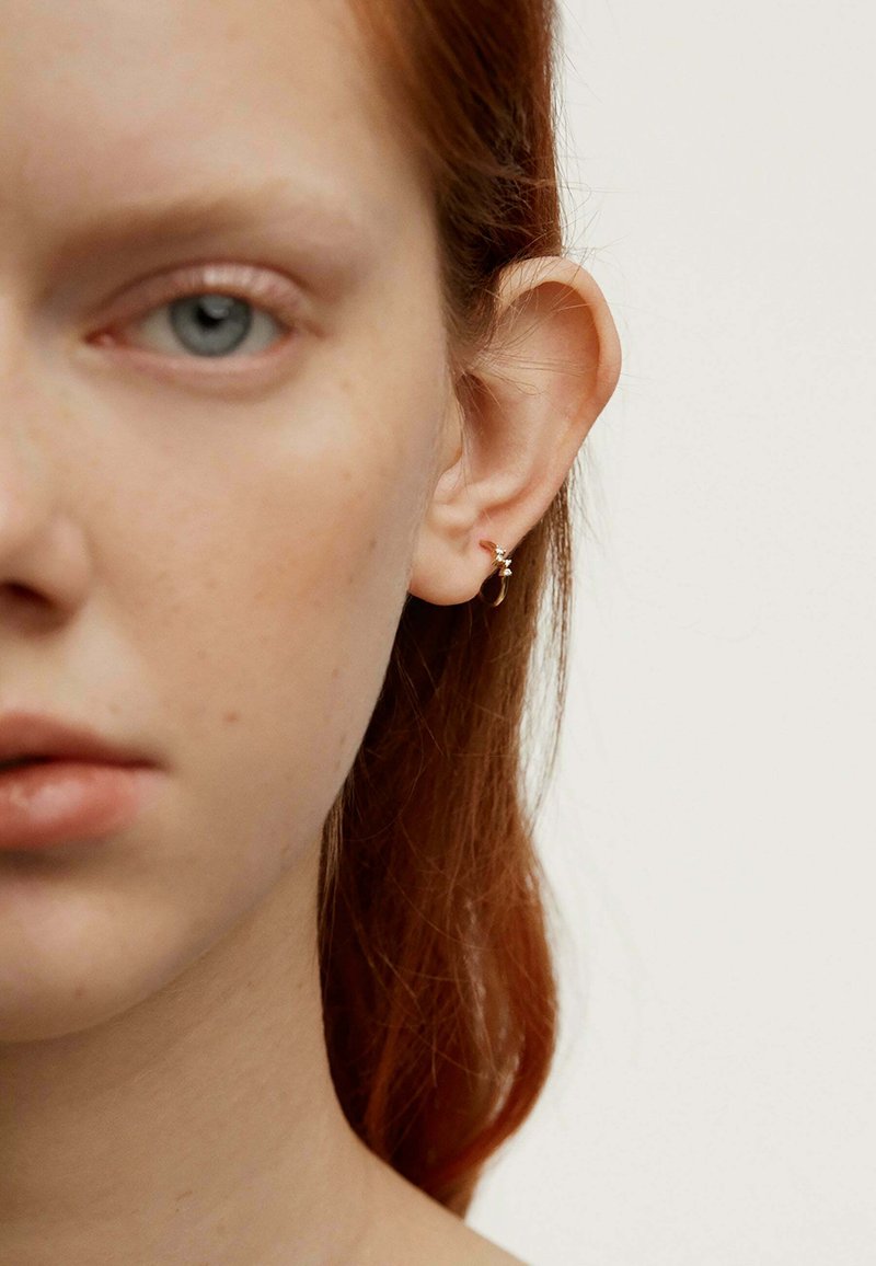 Pendiente de aro plateado con un pequeño acento de gema, llevado en el lóbulo, complementando la piel clara y el cabello rojizo. Primer plano de las características faciales.