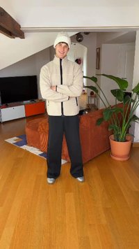 Svetlo bež flis jakna s črnimi poudarki, ohlapne črne hlače in srebrni športni copati. Notranja postavitev z lesenim tlemi in sobno rastlino v loncu.