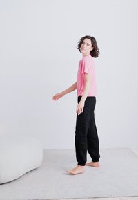 Pink short-sleeve t-shirt and black patterned sweatpants. Model stands barefoot on light carpet, with a light background and soft furniture nearby.
