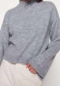 Szary sweter z dzianiny z wysokim kołnierzem i szczegółowym zamkiem na ćwierć. Posiada prążkowane mankiety i dół, lekko luźny krój oraz miękką teksturę.