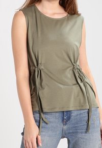 Olivgrön ärmlös topp med rund halsringning, rynkade sidodetaljer med justerbara band och en mjuk, len tygtextur.