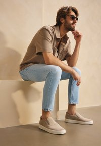 Béžové nazouvací boty se semišovým svrškem a bílou gumovou podrážkou. Světle modré upnuté džíny a hnědá košile s krátkým rukávem dotvářejí celý outfit.