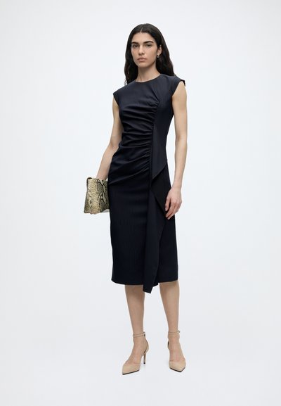 Femme portant une robe noire froncée sans manches, longueur genoux, tenant une pochette en peau de serpent et des chaussures à talons beiges, posant devant un fond uni.