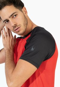 Camiseta deportiva roja y negra con manga corta, tela texturizada y un discreto logo de araña en el hombro izquierdo.