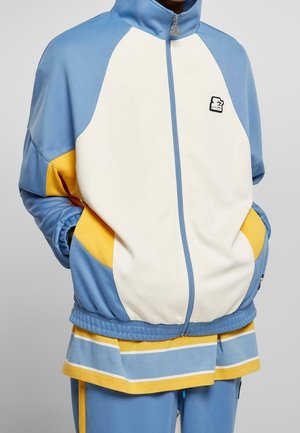 Person trägt eine blau-weiße-gelbe Jacke mit Reißverschluss, passende blaue Hose und darunter ein gestreiftes blau-gelbes Hemd.