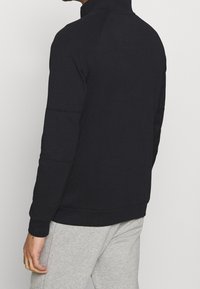 Muže, který má na sobě černou mikinu s dlouhým rukávem a světle šedé kalhoty, zobrazený zezadu na jednoduchém světlém pozadí.