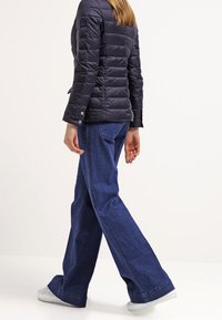 Kvinna i svart vadderad jacka, vida blå jeans och vita sneakers som går mot en enkel vit bakgrund.