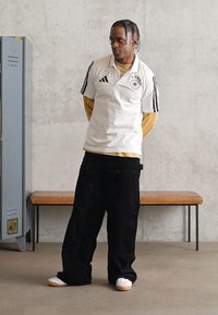 Homem com cabelo trançado a usar um jersey desportivo branco, uma camisa de mangas compridas amarela, calças largas pretas e ténis brancos, em pé ao lado de armários e um banco.