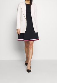 Lichtroze blazer over een zwarte jurk met een uitlopende rok en een horizontale gestreepte zoom in rood, wit en roze. Zwarte puntige hakken.