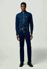 Camisa de mezclilla azul oscura con mangas largas, con un yugo de estilo western y un bolsillo; combinada con jeans oscuros de corte slim y botas de cuero negras hasta el tobillo.