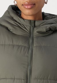 Giacca imbottita verde con cappuccio. Presenta una chiusura con zip, dettagli di cucitura e una texture liscia. Vista ravvicinata della parte superiore.