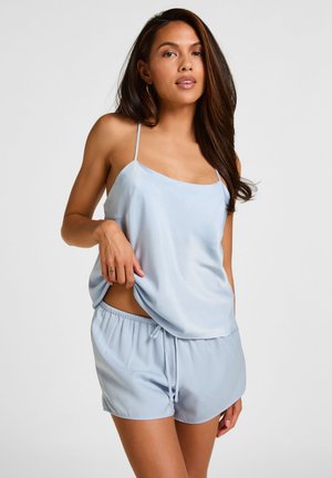 Frau mit langen dunklen Haaren, die ein hellblaues Satincamisole und passende Shorts trägt, vor einem schlichten weißen Hintergrund stehend.