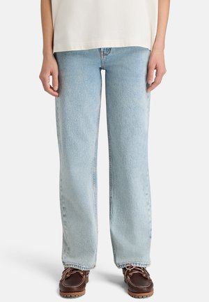 Persona che indossa jeans azzurri a gamba diritta, una maglietta bianca e scarpe marroni con lacci, in piedi su uno sfondo bianco semplice.