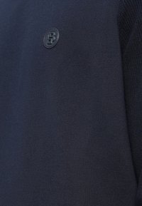 Jūras spēku džemperis, kas izgatavots no adītas auduma, ar mazu, apaļu melnu logotipu kreisajā krūtīs un ribotas detaļas uz pleciem.