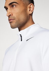 Pulover alb cu fermoar la jumătate, din material texturat, având logo-ul Nike negru pe piept. Dispune de un fermoar negru și un guler structurat.